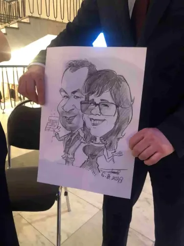Vtipná karikatúra páru z podujatia, kde muž drží v ruke pohár vína a žena má prehnane veľké oči a široký úsmev.